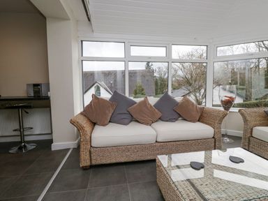 A seating area with wicker furniture and cushions by large windows at Whimhowe in Windermere