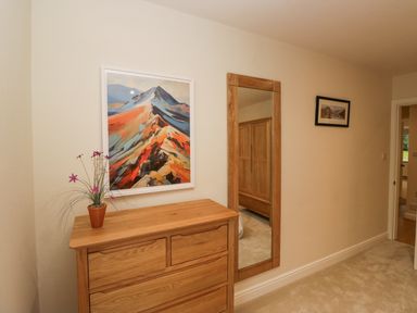 A wooden chest of drawers with a potted plant and mountain artwork above it next to a wall mirror and a framed picture at Whimhowe in Windermere