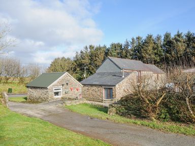 An outdoor scene with two stone buildings and a driveway surrounded by grass and trees at The Dairy Barn in Camelford