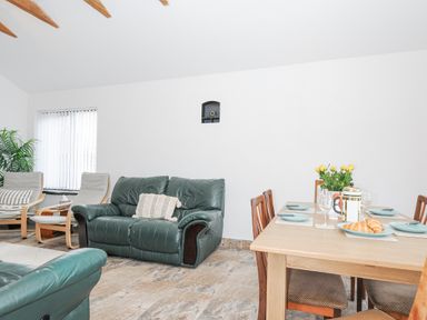 A living room with green leather sofas, wooden chairs, a dining table set with plates and croissants, and a plant by the window at The Dairy Barn in Camelford