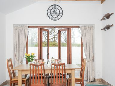 A dining room with a wooden table and six chairs near wooden folding doors with curtains at The Dairy Barn in Camelford