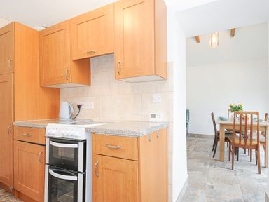 A kitchen with wooden cabinets and a white electric stove next to a dining area with a wooden table and chairs at The Dairy Barn in Camelford