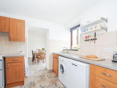 A kitchen with wooden cabinets, a washing machine, dishwasher, sink, knives on a magnetic strip, and a dining table with chairs visible in the next room at The Dairy Barn in Camelford