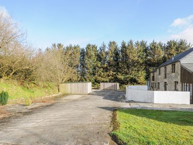 An outdoor area with a paved surface two wooden storage sheds a stone building and trees in the background at The Dairy Barn in Camelford