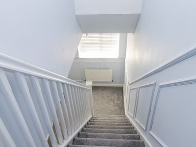 A carpeted staircase with white railing leading down to a landing with a radiator and window blinds at 9 Beckett Close in Colwyn Bay
