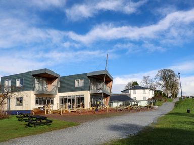 A modern building with balconies and a cafe sign next to a gravel path with picnic tables on a grassy area at Crathie Lodge in Banavie near Caol