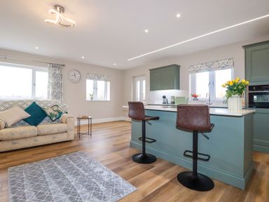 A kitchen with green cabinets and an island with two brown bar stools next to a living area with a beige sofa and a small table at Little Hillside in Redruth