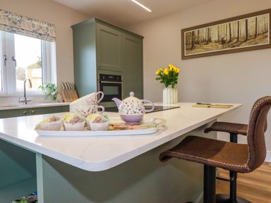 A kitchen with a white countertop island green cabinets two brown bar stools a teapot cups cupcakes and yellow flowers at Little Hillside in Redruth
