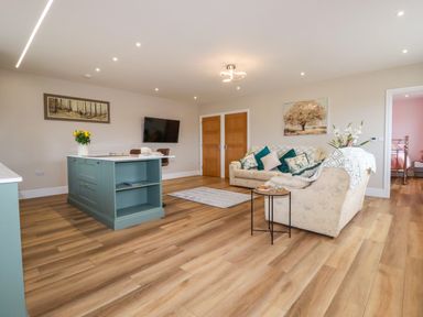 An open living room and kitchen area with a green kitchen island a sofa with cushions a small table a wall-mounted tv and wooden doors at Little Hillside in Redruth