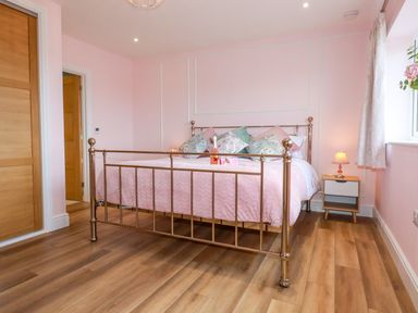 A bedroom with a metal bed frame, pink bedding, multiple pillows, a bedside table with a lamp, wooden floor, and pink walls at Little Hillside in Redruth