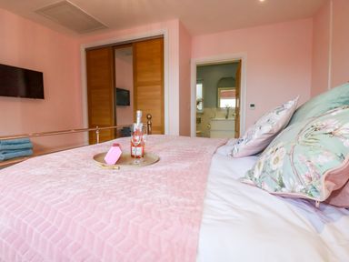 A bedroom with pink walls a bed with floral pillows a tray on the bed with a bottle and two glasses wooden closet doors and a wall-mounted TV at Little Hillside in Redruth