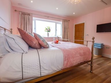 A bedroom with a metal frame bed pink bedding cushions a window with curtains a wooden door a wall mounted television and folded towels on a stool at Little Hillside in Redruth