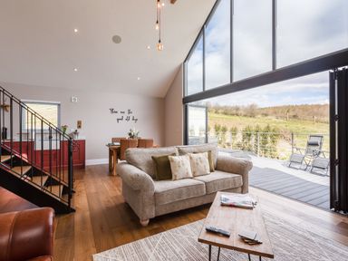 A living room with a beige sofa, coffee table, dining table, and large glass windows opening to a deck at Hare's Hill in Thornton-Le-Dale