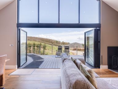 A living room with a sofa and pillows opening onto a deck with two chairs overlooking grass and a building at Hares Hill in Thornton-Le-Dale