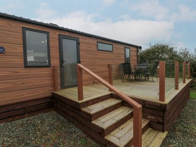 An exterior view of a wooden cabin with stairs leading to a deck with glass railing and outdoor table and chairs at Caldey Retreat in Hasguard Cross near Broad Haven