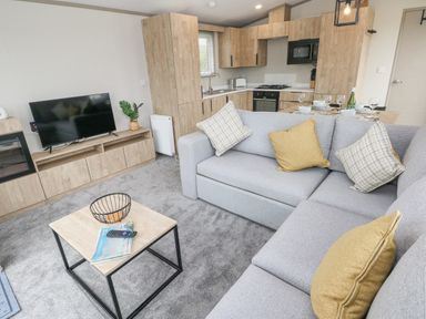 A living room with a grey sectional sofa and a wooden coffee table near a kitchen with wooden cabinets at Caldey Retreat in Hasguard Cross near Broad Haven