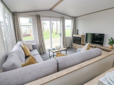 A living room with grey sofas, a coffee table, a TV on a wooden stand, and large windows with checkered curtains at Caldey Retreat Hasguard Cross near Broad Haven