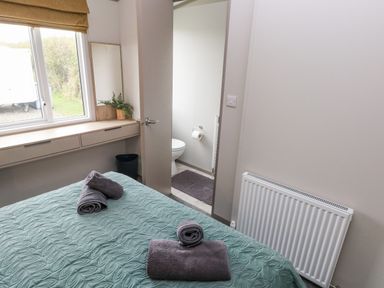 A bedroom with a bed covered by a green quilt and towels on top a window with a blind a doorway leading to a bathroom with a toilet visible a mirror and a plant on a shelf Caldey Retreat Hasguard Cross near Broad Haven