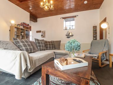 A living room with a gray sectional sofa and pillows a wooden coffee table with a tray and flowers a gray chair with a blanket a wooden ceiling and white walls with a small window at Owl Cottage in Ashen Cross Pelynt