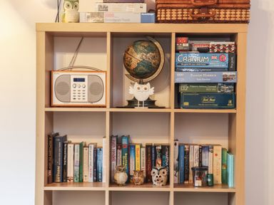 A wooden shelving unit with books, board games, a globe, a radio, owl figurines, and a wicker basket at Owl Cottage in Ashen Cross Pelynt