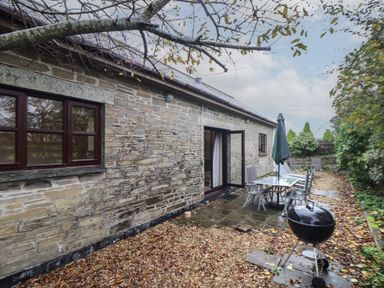 An outdoor patio area with a glass table and chairs next to a stone building with open doors and a barbecue grill surrounded by trees at Owl Cottage in Ashen Cross Pelynt