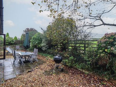 A garden patio with a glass table and chairs a closed umbrella a barbecue grill and surrounding plants at Owl Cottage in Ashen Cross Pelynt