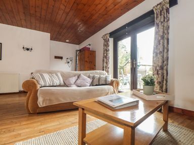 A living room with a sofa, cushions, wooden coffee table with a plant, wooden floor, and glass doors with floral curtains at Court Cottage in Ashen Cross, Pelynt