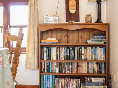 A wooden bookshelf with books, puzzles, a radio, and decorative items next to a window with curtains at Court Cottage in Ashen Cross, Pelynt