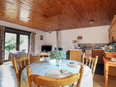 A dining table with chairs and flowers in a room with a kitchen and seating area at Court Cottage in Ashen Cross Pelynt