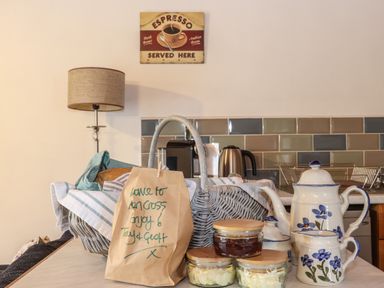 A kitchen counter with a wicker basket, a brown paper bag with a note, glass jars with lids, a floral ceramic teapot set, a lamp, and a tiled backsplash at Court Cottage in Ashen Cross Pelynt