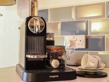 A coffee machine with coffee capsules in a jar and stacked cups and saucers in a kitchen at Court Cottage Ashen Cross Pelynt