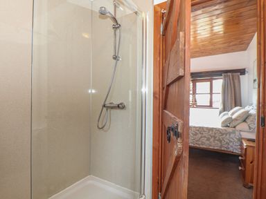 A shower enclosure with glass walls next to a wooden door leading to a bedroom with a bed and window at Court Cottage Ashen Cross Pelynt