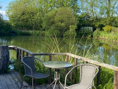 An outdoor seating area with a small glass table and two chairs on a wooden deck by a pond with trees in the background at Court Cottage in Ashen Cross Pelynt