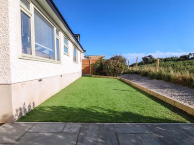 A side garden with artificial grass next to a house and a gravel path with a wooden fence at Awel-Y-Mor in Trearddur Bay
