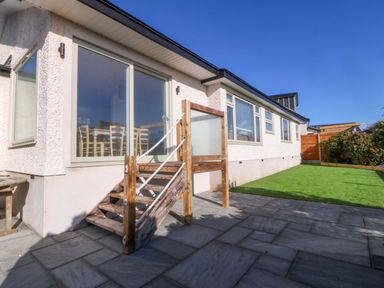 A patio with stone tiles and wooden steps leading to sliding glass doors next to a lawn at Awel-Y-Mor in Trearddur Bay