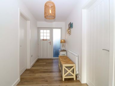 A hallway with wooden flooring a white entrance door a bench with baskets and a lamp on a side table at Awel-Y-Mor Trearddur Bay