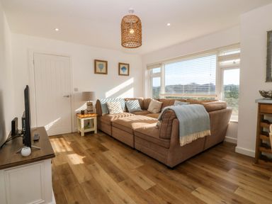 A living room with a brown sectional sofa near a window with blinds and a television on a stand at Awel-Y-Mor in Trearddur Bay