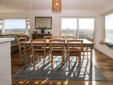 A dining room with a wooden table and eight chairs on a rug with a sideboard and windows at Awel-Y-Mor in Trearddur Bay