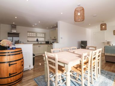 A dining area with a wooden table and chairs next to an open kitchen with cabinets and appliances at Awel-Y-Mor in Trearddur Bay