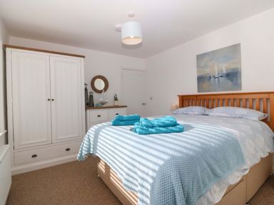 A bedroom with a wooden bed, light blue bedding, turquoise towels on the bed, a white wardrobe, a dresser with decorative items, and a sailboat painting on the wall at Awel-Y-Mor in Trearddur Bay