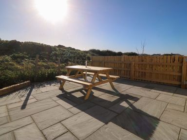 A patio with a wooden picnic table and fence with bushes in the background at Awel-Y-Mor in Trearddur Bay
