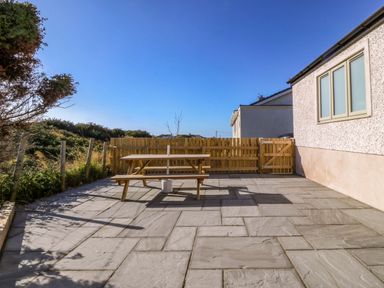 A fenced patio area with a wooden picnic table and bushes at Awel-Y-Mor in Trearddur Bay
