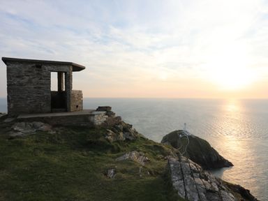 A stone shelter on a grassy cliff overlooking a lighthouse on a promontory at sunset at Awel-Y-Mor in Trearddur Bay