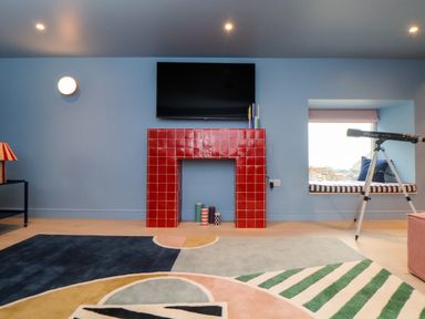 A room with a red tiled fireplace a wall mounted TV a telescope next to a window seat with cushions and a colorful geometric rug on the floor at Mavericks Wave in St Agnes