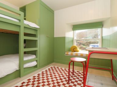 A bedroom with green bunk beds a green wardrobe a window seat a red stool a desk and a patterned rug at Mavericks Wave in St Agnes