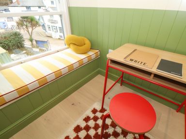 A small workspace with a red stool desk with notebooks and a striped bench seat with yellow pillow by a window at Mavericks Wave in St Agnes