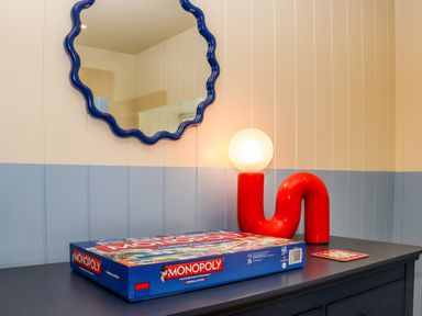 A blue dresser with a monopoly board game a red lamp with a round white bulb and a blue wavy edged mirror on the wall at Mavericks Wave in St Agnes