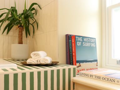 A corner with a potted plant, rolled white towels on a tray, and books on a windowsill at Mavericks Wave in St Agnes