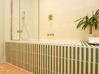 A bathroom with a bathtub that has green and white vertical striped tiles and a glass shower screen with a plant and folded towels on the side at Mavericks Wave in St Agnes