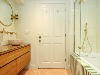 A bathroom with a wooden vanity cabinet and round mirror next to a white door and a bathtub with green and white tiled surround at Mavericks Wave in St Agnes